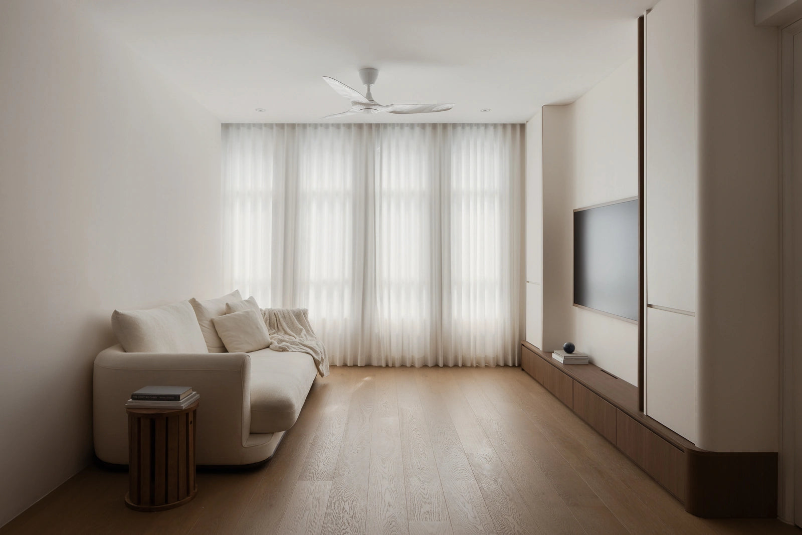 Minimalist living room featuring a cream sectional sofa, curved TV feature wall, light wood flooring, and sheer day curtains.