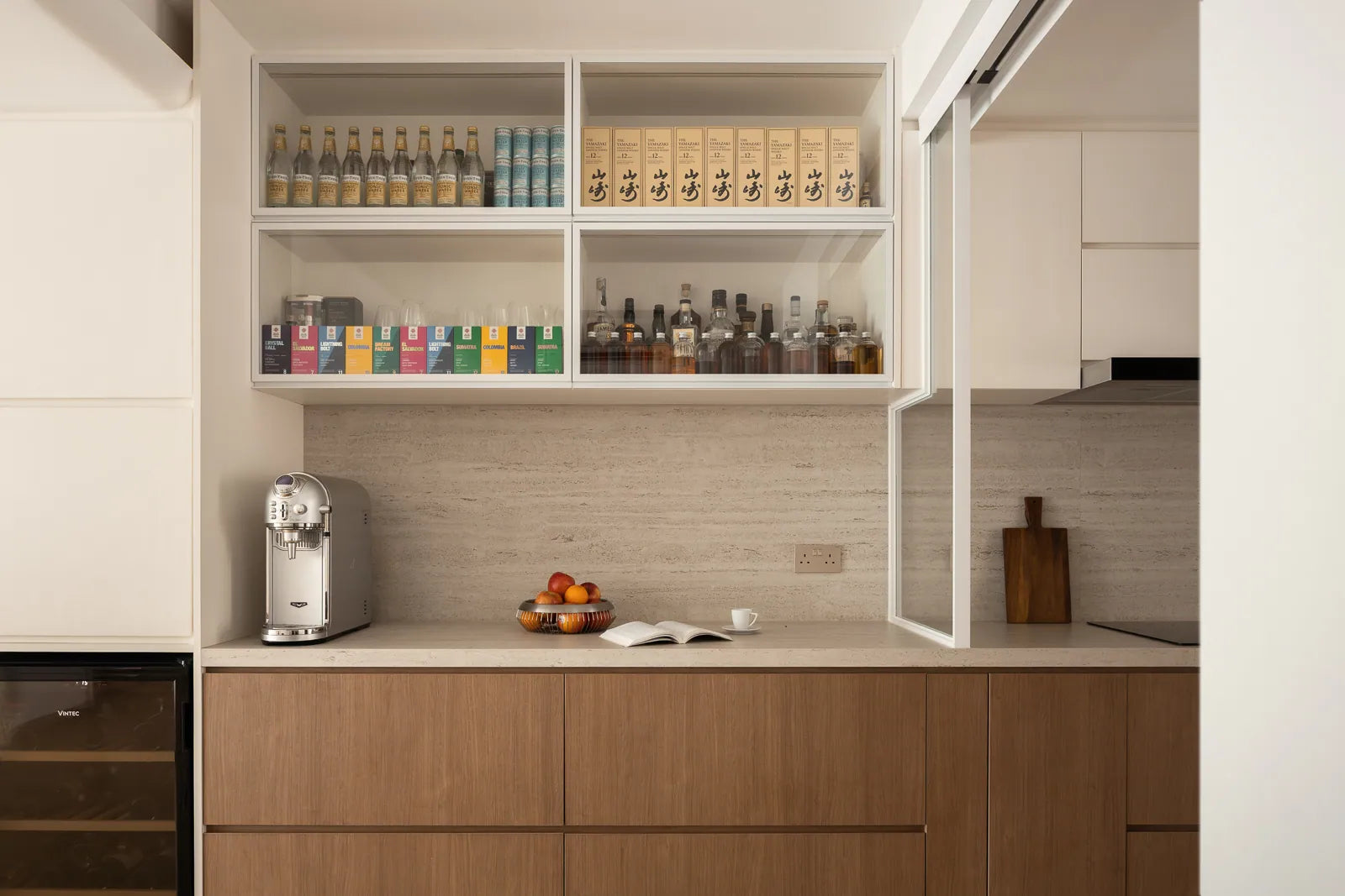 Minimalist dry kitchen with a glass display cabinet, travertine-inspired backsplash, and warm wood cabinetry