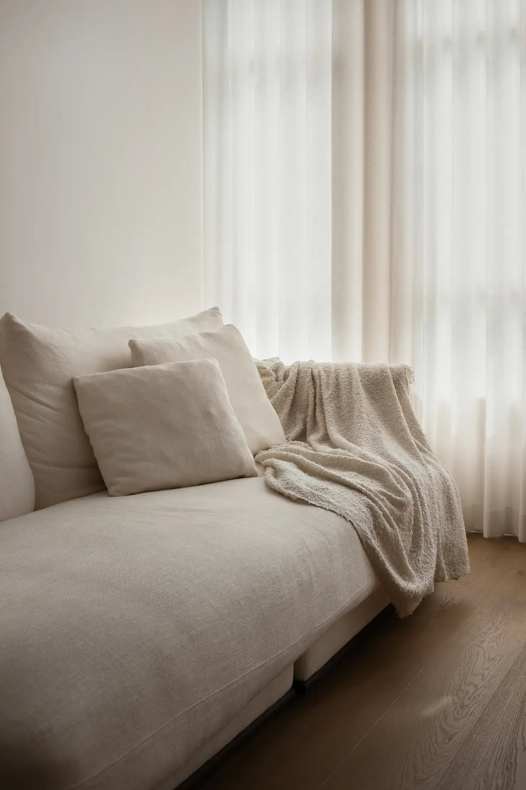 Cream sectional sofa with cushions and a throw in a bright minimalist living room with sheer day curtains.