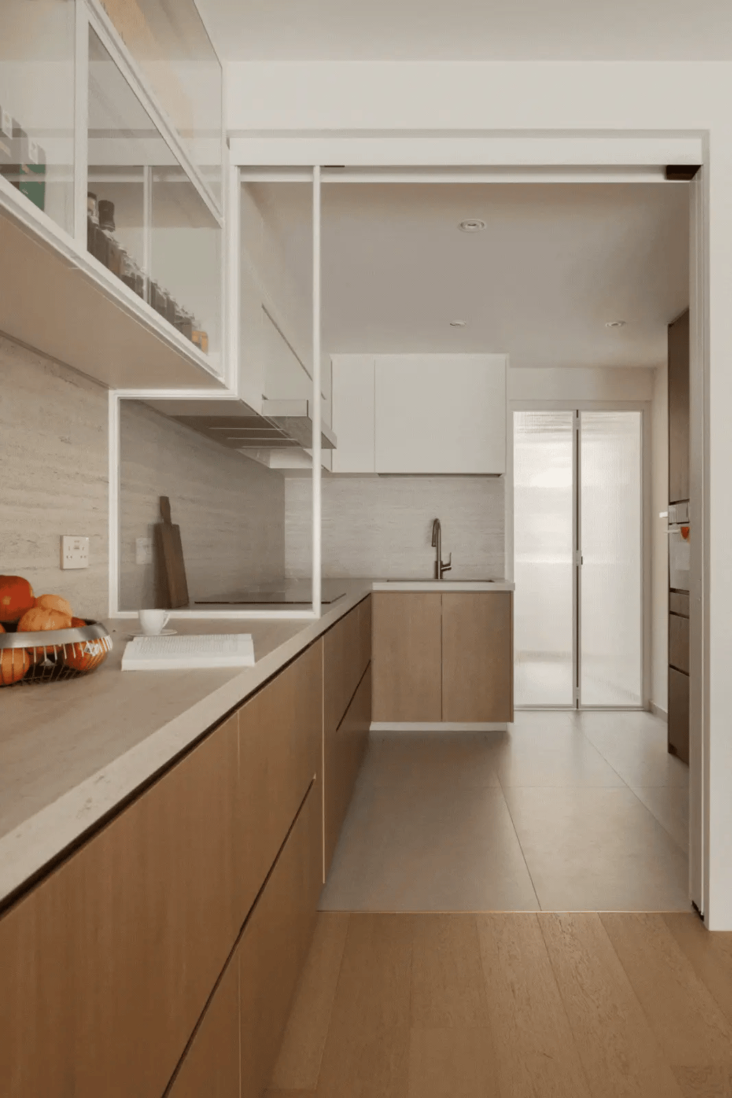Minimalist kitchen with a glass display cabinet, travertine-inspired backsplash, and warm wood cabinetry.