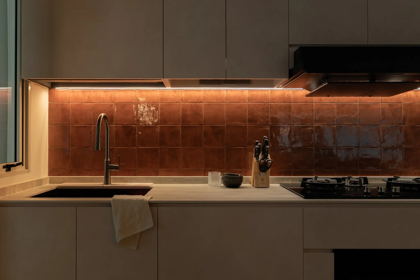 Modern kitchen with terracotta-toned square tile backsplash, cream cabinetry, a black sink and under-cabinet lighting